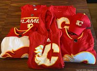 Six Calgary Flames fan wear items including caps, t-shirts, hockey jerseys, and a hoodie, showing front views laid flat on wooden background.