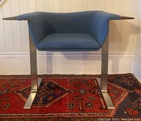 Front view of mid century chair showing blue upholstered seat, walnut arm rests and stainless steel legs on a patterned rug.