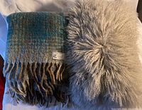 Blue/tan plaid Piuma wool/polyamide blanket folded next to fluffy grey Mongolian lamb pillow on a light surface.