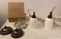 Two porcelain pendant lights with white unglazed shades and antiqued brass lamp holders laid out with mounting plates and ceiling roses on white cloth surface, with a cardboard box in background.