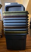 Stacked lot of nine Rubbermaid Roughneck 68 litre storage bins of various colors including dark blue, light blue, green, and brown lids in need of cleaning.