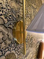 Close side view of one unlacquered brass wall sconce mounted on decorative wallpaper showing circular mounting plate and brass rod with cane wrap detail