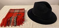 Photo of navy wool fedora hat and folded red, black, white plaid scarf with fringes on white cloth
