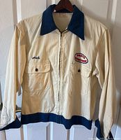 Front view of cream work jacket with navy collar, zipper front, two button flap pockets, "Pete" name embroidery left chest, "Parts Department" patch right chest.