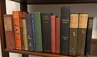 Shelf display of 13 vintage/antique hardcover books in various bindings and colors including leather bound titles.