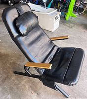 Side angle view of the black leather style reclining chair showing the metal frame, wooden armrests, and attached pillow.