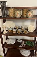 Wide angle photo showing all items on shelves including floral decorated metal tins, white porcelain plates, child's tea set with painted figures, white porcelain tureens, ceramic candleholder, and metal candleholder.