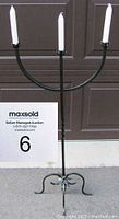 Full front view of the black metal standing candle holder with three white taper candles inserted, displayed against a brown garage door.