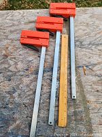 Three 16 inch metal bar clamps with orange plastic lever handles placed on a wooden surface next to a 12 inch ruler for scale.