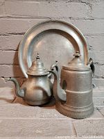 Wide shot of the antique pewter charger plate, tea pot, and coffee pot against a brick background