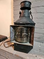 Full view of the antique ship's lantern showing the metal top with ventilation holes, hanging handles, convex glass lens in a metal frame, and attached electrical cord.