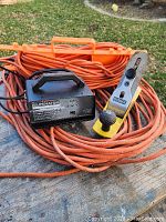 Photo showing Motomaster battery charger, orange extension cords, and Narex wood planer together on a wooden surface outside.