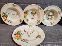 Four decorative china plates arranged in two rows: top row has three plates with different stencilled images/phrases, bottom row has one plate with steer skull. All have floral patterns and gold trim.
