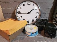 Photo showing quartz wall clock, box of manila envelopes, electric pencil sharpener, and stack of DVD-R discs on a grey surface.