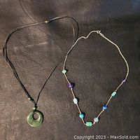 Pair of necklaces laid flat on dark surface, showing the jade pendant necklace and the silver chain with gemstones necklace.