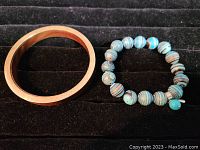 Pair of bracelets on black fabric background. Left is a smooth mahogany wood bangle with brass inside. Right is a bracelet with large striped glass beads in shades of blue and white.