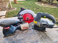 Overview of three power tools on wooden surface outdoors: a Ryobi circular saw, a Ryobi sheet sander, and a Pro-Use jigsaw