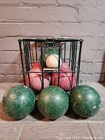 Green and red bocce balls along with one white pallino ball arranged inside and outside the metal carrying cage.