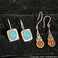 Two pairs of earrings displayed side by side on black fabric. One pair has blue turquoise stones, the other has amber in Celtic silver setting.