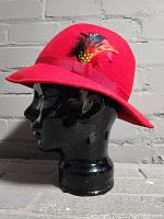 Side angle view of vintage red felt hat showing brim, hatband, and feather decoration.
