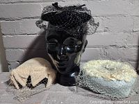 Three vintage hats arranged together. The black hat is placed on a black mannequin head, flanked by the other two hats: cream and blue with floral texture, each featuring netting.