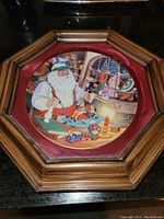 Framed Christmas plate showing Santa Claus painting toys, with a snowy window in the background, mounted in an octagonal wooden frame with reddish matting.