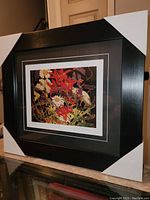 Front view of framed art print showing vibrant flower abstract artwork, black wood frame with protective corner covers.