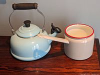 Photo of light blue enamel kettle with wooden handle and black lid knob alongside beige enamel pot with red rim and handle on wooden surface.
