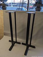 Pair of black metal speaker stands photographed against a white backdrop showing full height and structure.