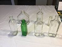 Full view of 7 antique glass bottles of various shapes and one green bottle on white cloth surface