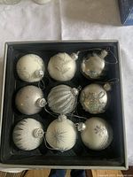 Set of 9 assorted silver and white glass Christmas ball ornaments displayed in black box with varied patterns including glitter stripes, snowflakes, swirls, and textured finishes.