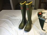 Front view of the pair of tall olive drab rubber boots with yellow trim, branded Lacrosse.
