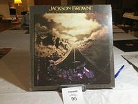 Front cover of Jackson Browne LP album 'Running on Empty' showing drum set on road and cloudy sky artwork.