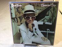 Front cover of Elton John's Greatest Hits LP showing Elton John in white suit sitting at a piano.