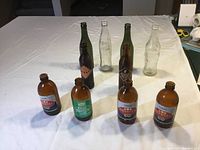 Front view of eight bottles arranged on white surface showing clear Coca-Cola bottles, dark green beer bottles and brown stubby bottles with labels