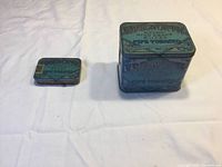 Photo showing both Edgeworth pipe tobacco tins, one small pocket-sized and one larger storage tin on a light-colored fabric background.