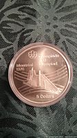 Obverse side of coin showing Montreal Olympic stadium design with Olympic rings, Montreal 1976, Olympiade XXI Olympiad, and 5 Dollars denomination.