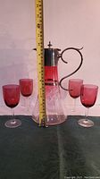 Photo showing carafe with measuring tape indicating height around 13 inches with 4 cranberry glass goblets arranged around it.