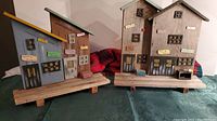 Photo of two driftwood fishing village models on wooden bases, showing rustic building facades with multiple windows, doors, and small colorful signs.