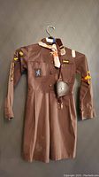 Brown Girl Guides of Canada uniform dress displayed on hanger, showing full front with badges and patches on sleeves.
