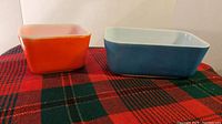 Two vintage Pyrex butter or condiment dishes, one red-orange and one blue, displayed on a plaid cloth.