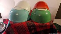 Overview of four vintage Pyrex mixing bowls in red, green, blue, and white stacked on red and black plaid cloth.