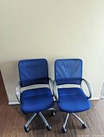 Two blue office chairs with mesh backs and silver armrests sitting on a wooden floor against a beige wall.