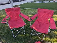 Two red folding camping chairs set up outdoors on grass, showing fabric seats, armrests with cup holders, and metal legs.