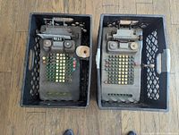 Top view of two Victor adding machines in black crates showing keys, levers, and paper roll on left machine.
