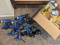 Seven cordless drills laid on carpet floor next to open box with spotlights in packaging