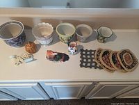 Overview of all items in the lot spread out on a white countertop showing seven decorative pots of different shapes and colors, three figurines (pumpkin, rooster, bird), a white metal decorative bracket, and four coasters with a holder.