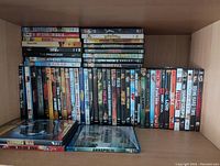 DVD box lot placed on wooden shelf, showing various DVD titles arranged upright and stacked horizontally, displaying a variety of movie genres.