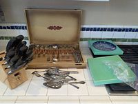Full view of all items on the counter including knife block, flatware case with utensils, loose flatware pieces, and display plates in boxes.