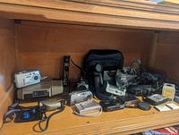 Wide shot showing multiple digital compact cameras, camcorder, chargers, camera grips, bags, and accessories placed on wooden shelf